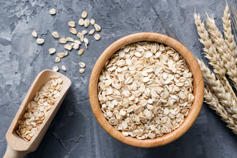 rolled oats in wooden bowl and golden wheat ears on stone background